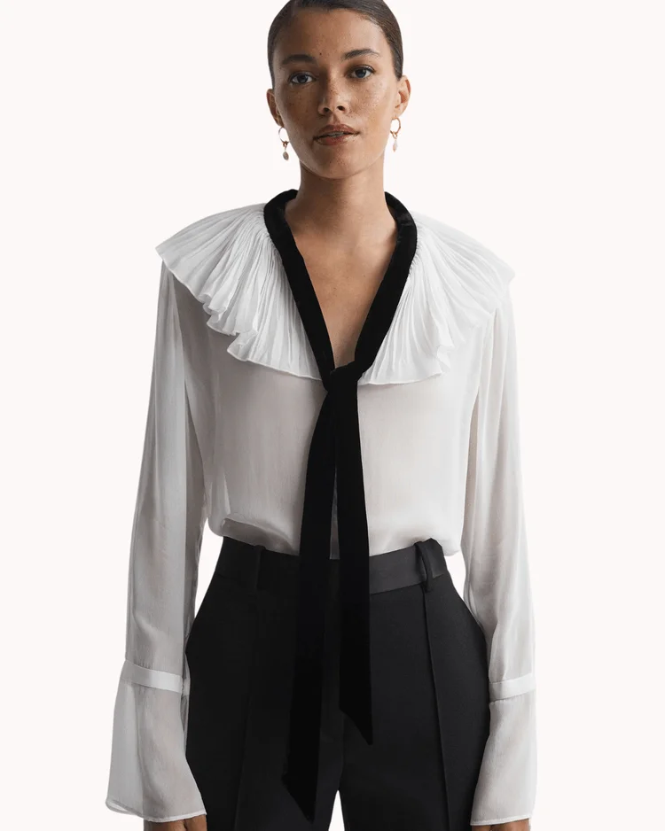 Model poses wearing ruffle neck sheer blouse with bow collar