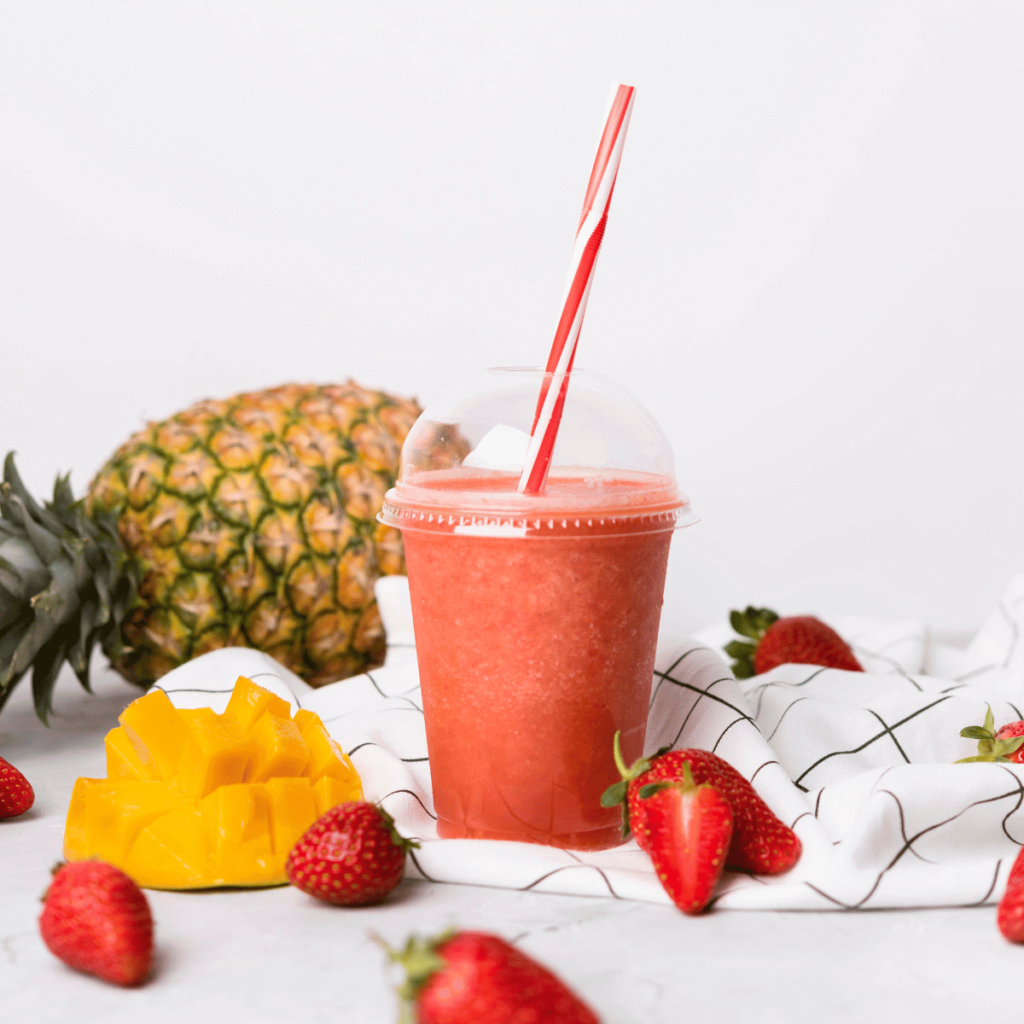 Upclose shot of smoothie filled lidded cup surrounded by pineapple, strawberries and a mango. 9 Ways To Stay Hydrated (That Isn’t Water)