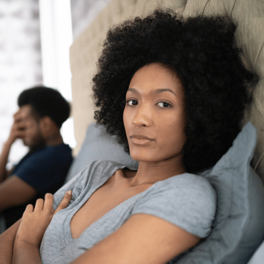 Woman with afro looks fed up she is sitting up beside her partner in bed. Wounded feminine energy