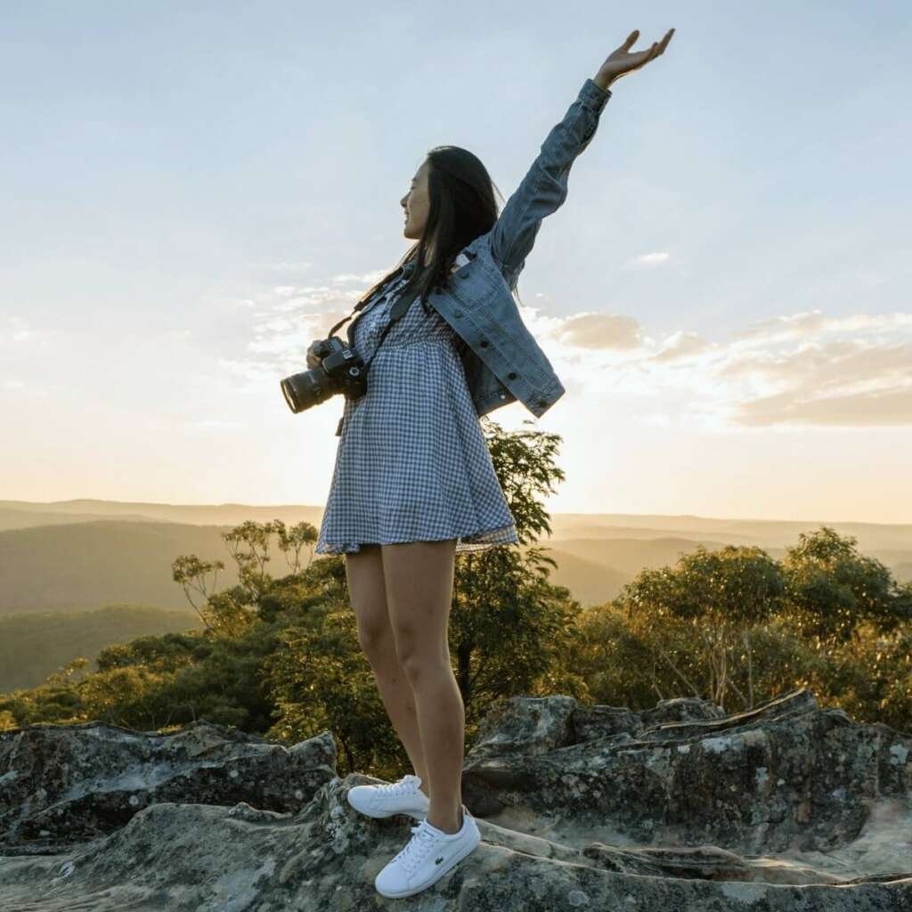 Medium shot of woman with a camera around her neck outside on a mountain top surrounded by trees. Self-care Sundays