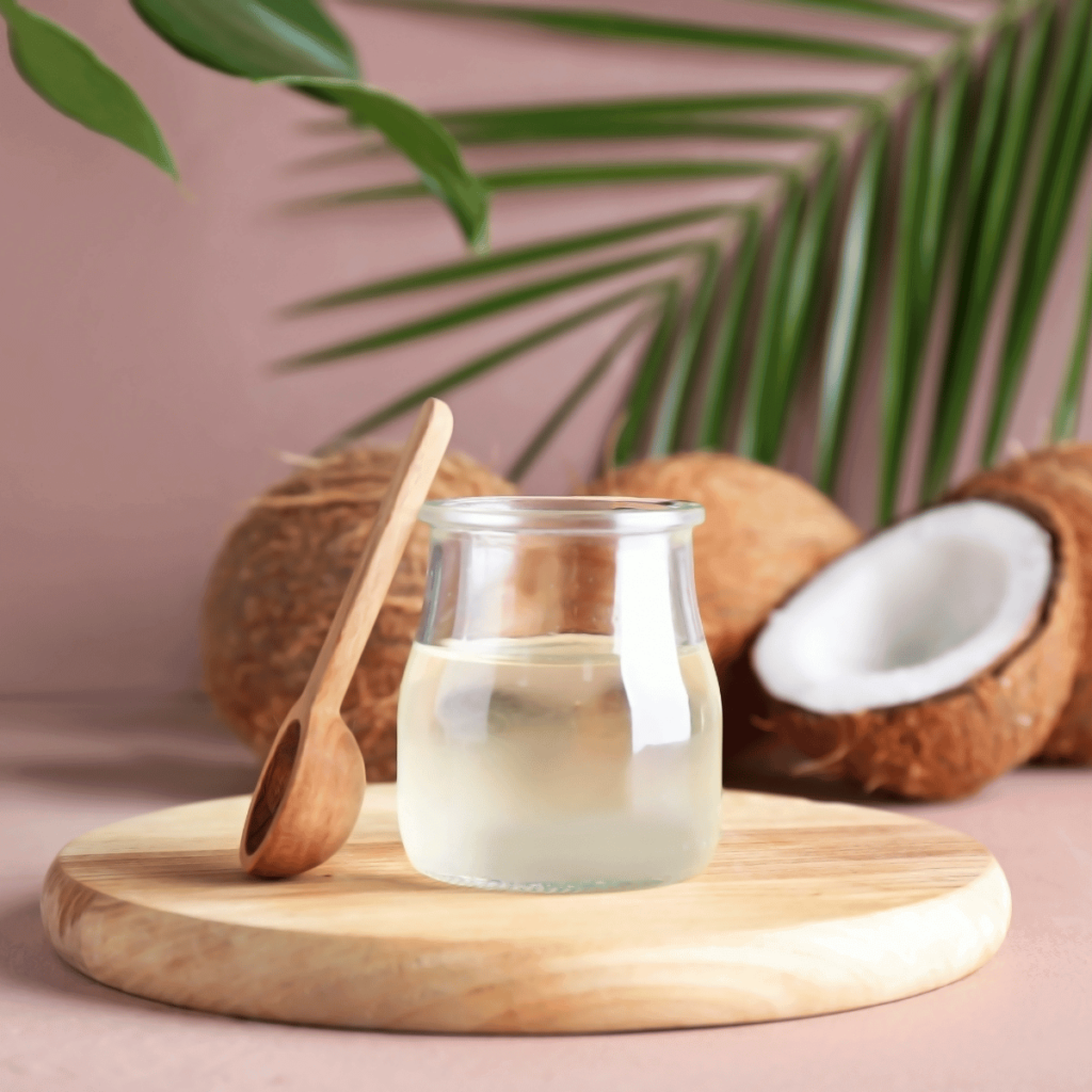 A glass of coconut oil on a wooden board surrounding by coconuts and leaves