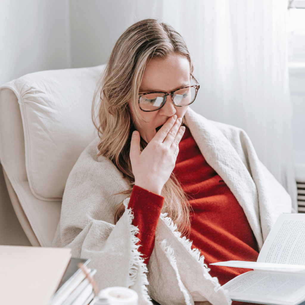 Woman with glasses yawning while sitting in a chair with a book in her hand