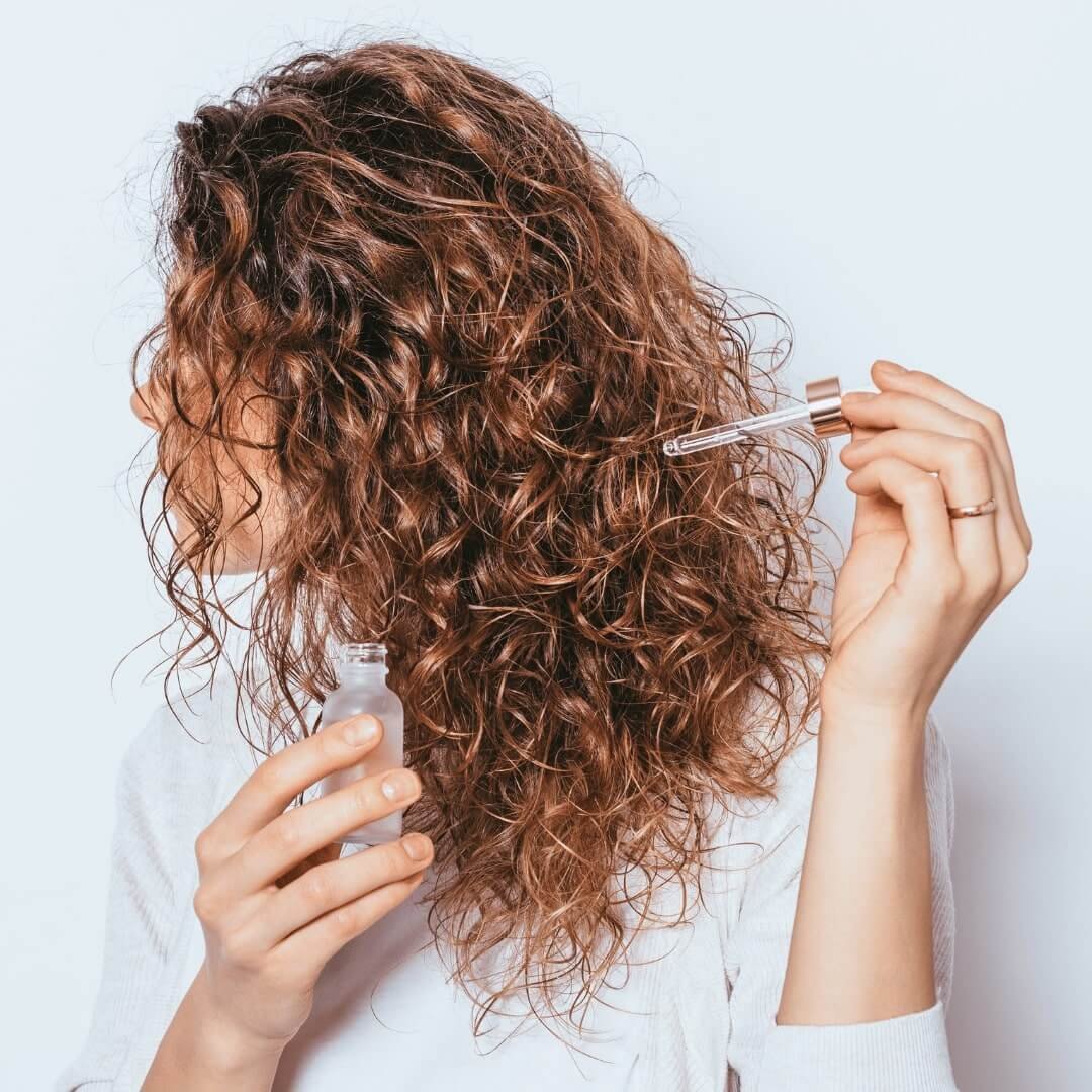 side view of woman using hair serum to her curly hair. Argan oil vs jojoba oil